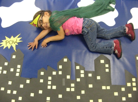 CCS Home Page Photo of CCS child, dressed up as a superhero and laying on a floor poster to create the illusion of flying over buildings..