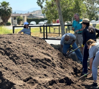 50% of the photo is a large pile of compost in the left corner. On the right side of the image is 4 people shoveling compost, 2 are bending over. One woman is standing behind the pile in a white hat looking on to the pile.