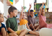 Group of small nursery school children sitting on floor indoors in classroom, raising hands. Childcare/Preschool Staff and Volunteers